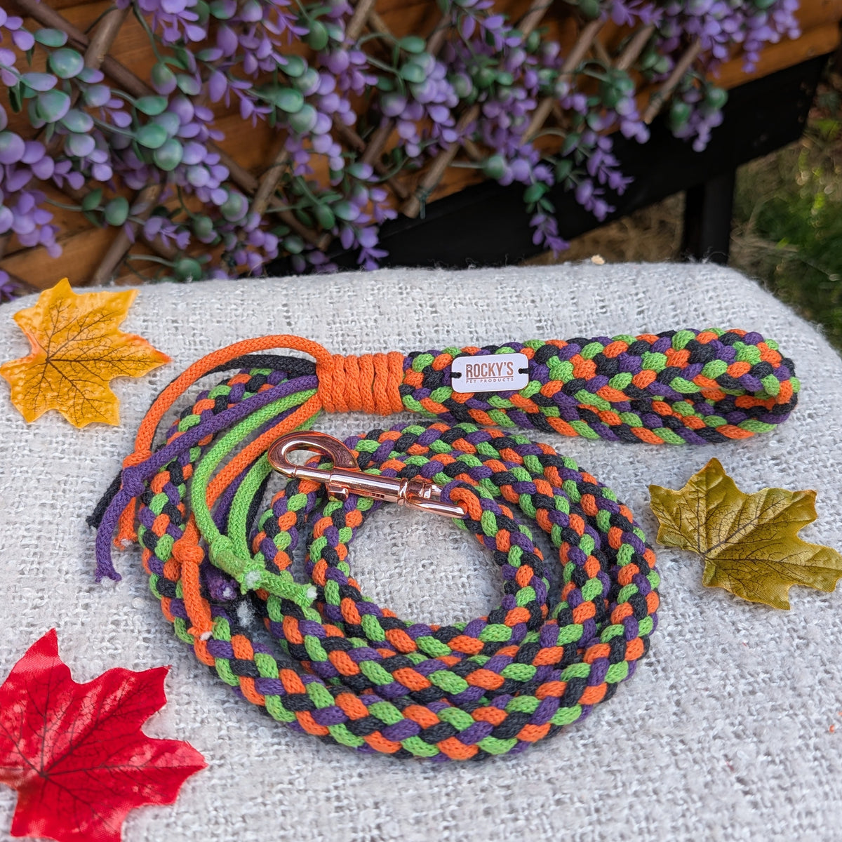 A braided dog lead with orange, green, and black cords and a metal lead hook displayed on a textured white fabric surface with decorative autumn leaves and a purple floral background.