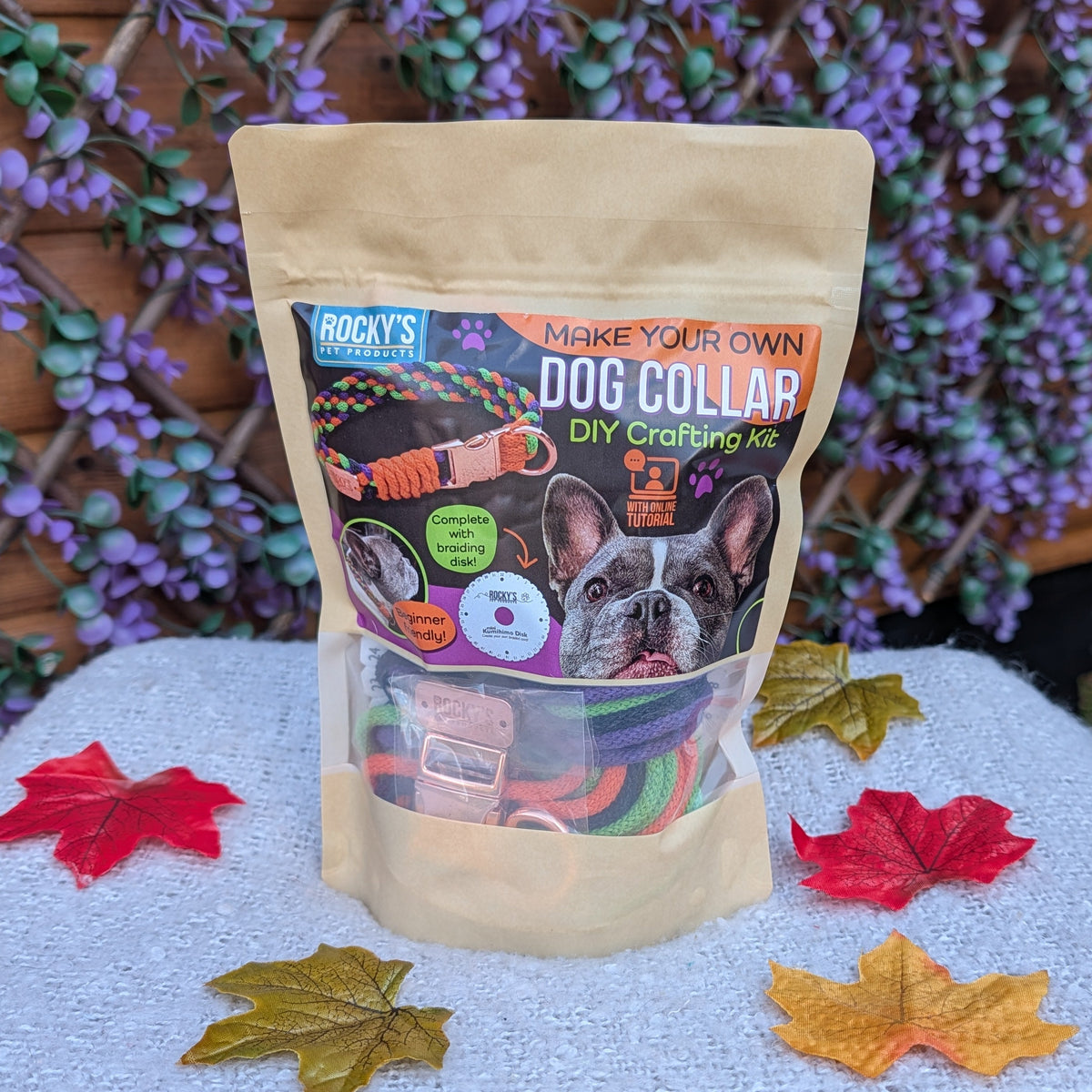 A stand-up pouch of Rocky's DIY Dog Collar supplies sits on a white textured fabric surface outdoors, with decorative autumn leaves around it and a wooden fence with purple flowers in the background.
