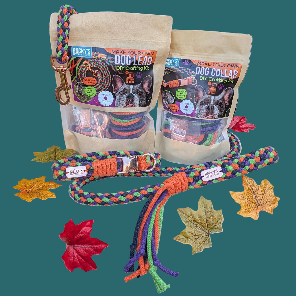 Two bags of DIY craft kits, dog leashes and collars displayed on a white textured blanket outdoors, with a colorful braided leash laid out in front and decorative autumn leaves on a wooden fence background.
