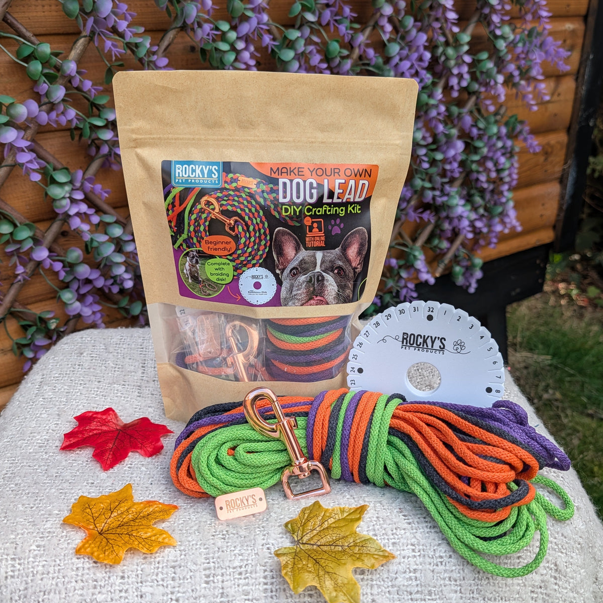 A dog lead craft kit by Rocky's Pet Products on a light gray fabric-covered stool in front of a wooden fence with purple flowers. The kit includes a tan stand-up pouch, a coiled multicolor rope lead, a white plastic agility jump, and decorative autumn leaves.
