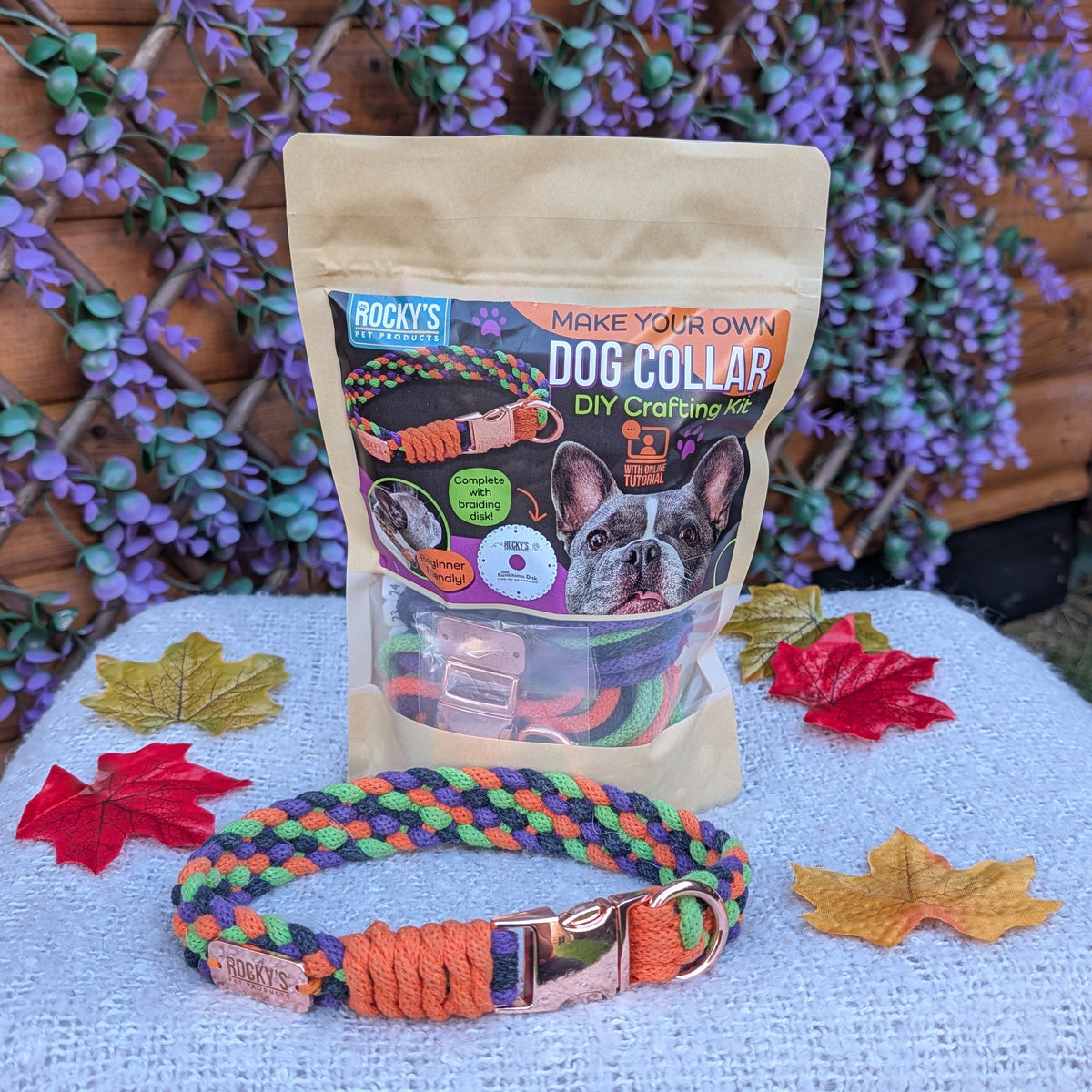 A bag of Rocky's Pet Products Dog Collar DIY Kit make your own dog collar sits on a white textured blanket outdoors, with a colorful braided dog collar placed in front. The background features a wooden fence decorated with purple flowers.