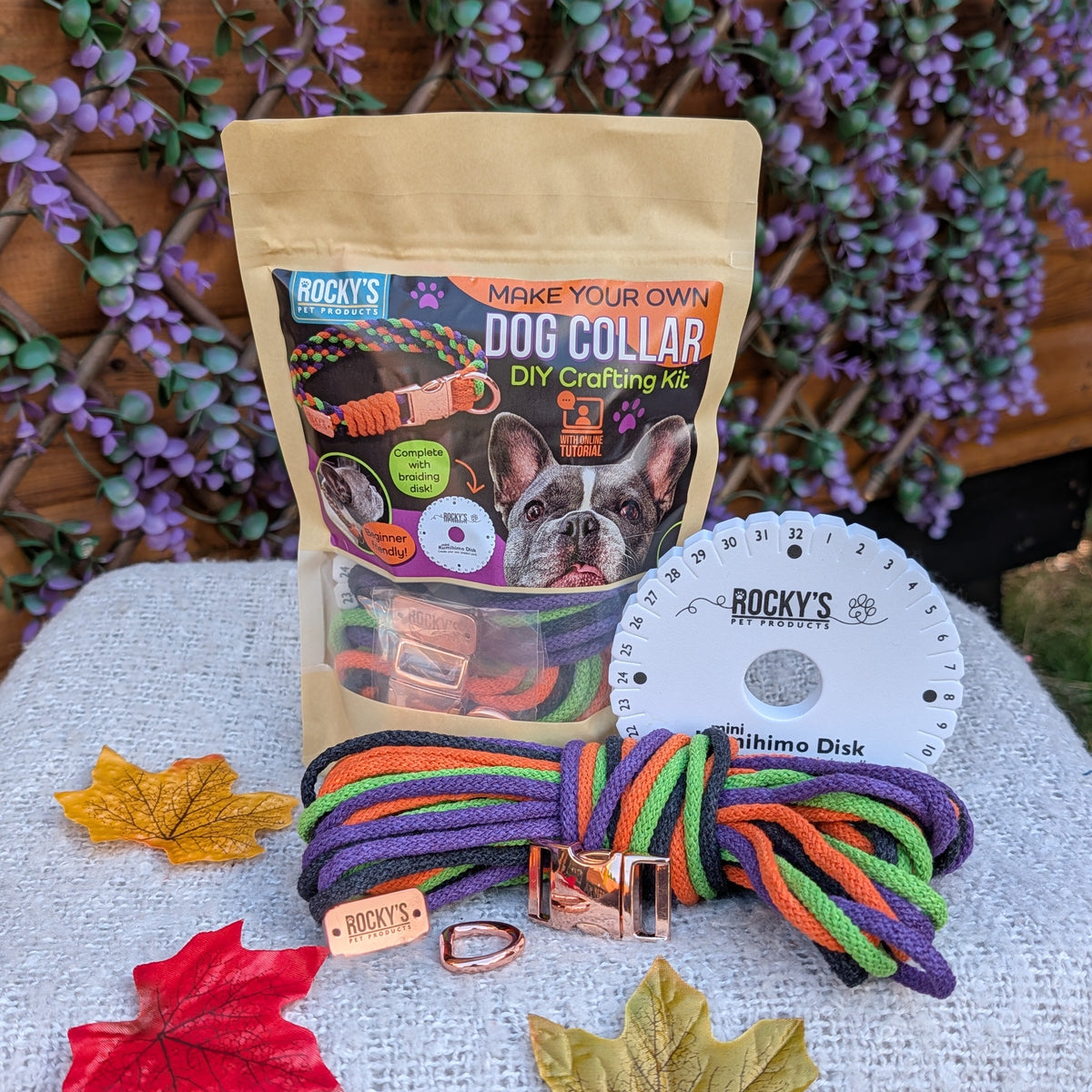 A dog collar kit by Rocky's displayed on a light gray fabric-covered table. The kit includes a tan stand-up pouch labeled 'Rocky's Make Your Own Dog Collar,' a white circular plastic spool, and a knotted rainbow-colored rope collar with a copper-toned metal buckle.