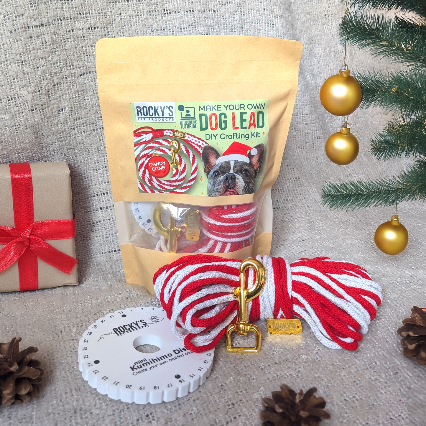 Dog lead DIY crafting kit with packaging, red and white rope, and gold clip on a textured surface with Christmas decorations.