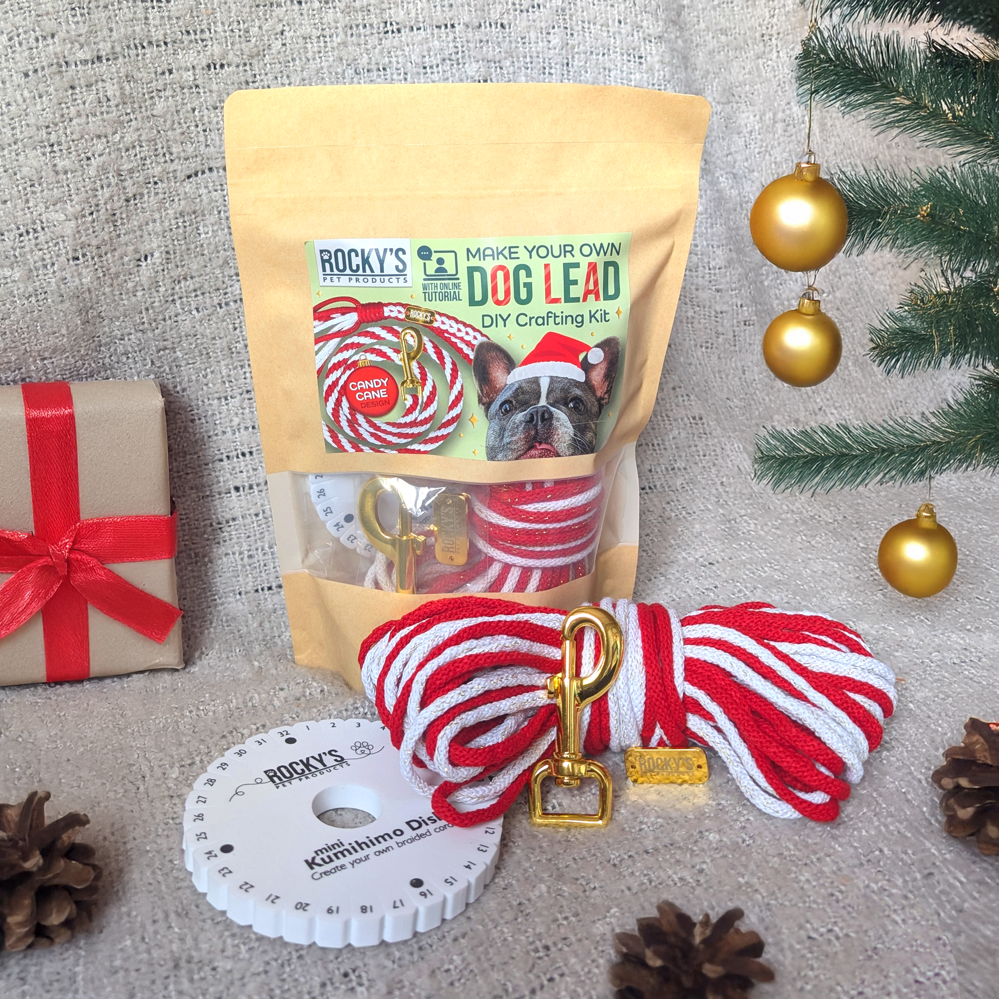 Dog lead DIY crafting kit with packaging, red and white rope, and gold clip on a textured surface with Christmas decorations.